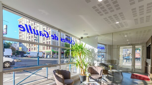Un moderno vestíbulo de hotel con ventanas amplias, un mostrador de recepción, asientos cómodos y una planta en maceta.