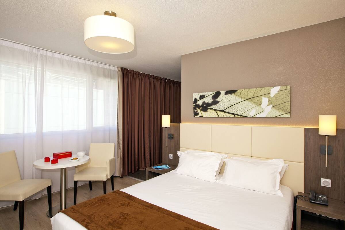 A hotel room with a bed, a desk with a chair, a small table with a red box, a phone, and a painting on the wall.