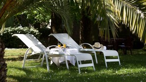A sunlit outdoor seating area with white lounge chairs and a table, surrounded by lush greenery and palm trees.