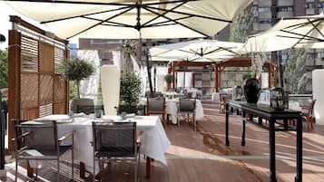 Un espace extérieur aménagé pour les repas, avec des tables et des chaises, protégé par de grands parasols et entouré de clôtures en bois et de plantes.