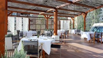 Un patio al aire libre con mesas y sillas de madera, cubierto por una pérgola de madera, con vistas a una zona residencial.