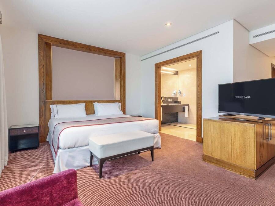 A hotel room with a large bed, a TV, a wooden cabinet, and a bathroom visible through an open door.