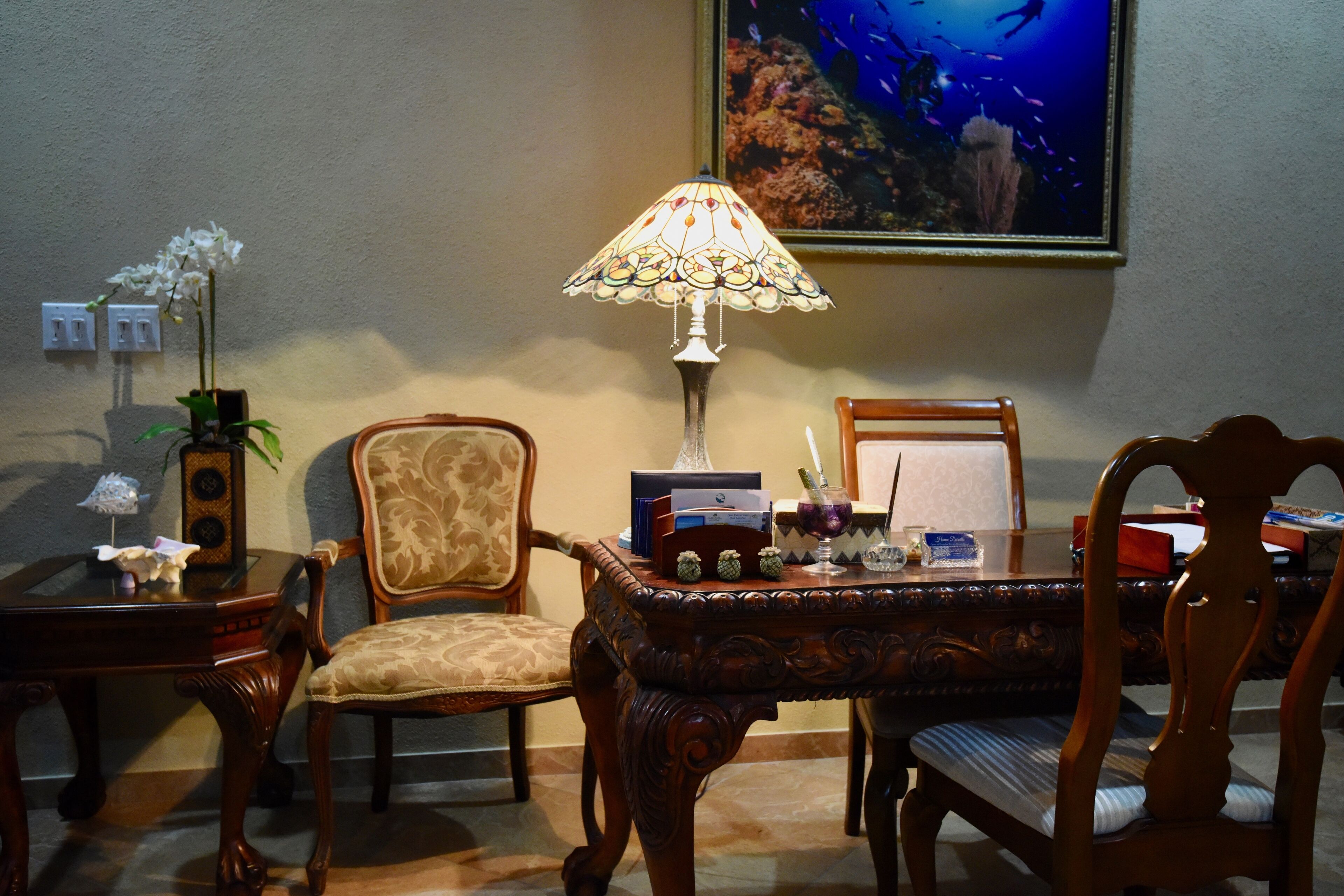 A room with a wooden table, two chairs, a lamp, a framed picture, and a potted plant.