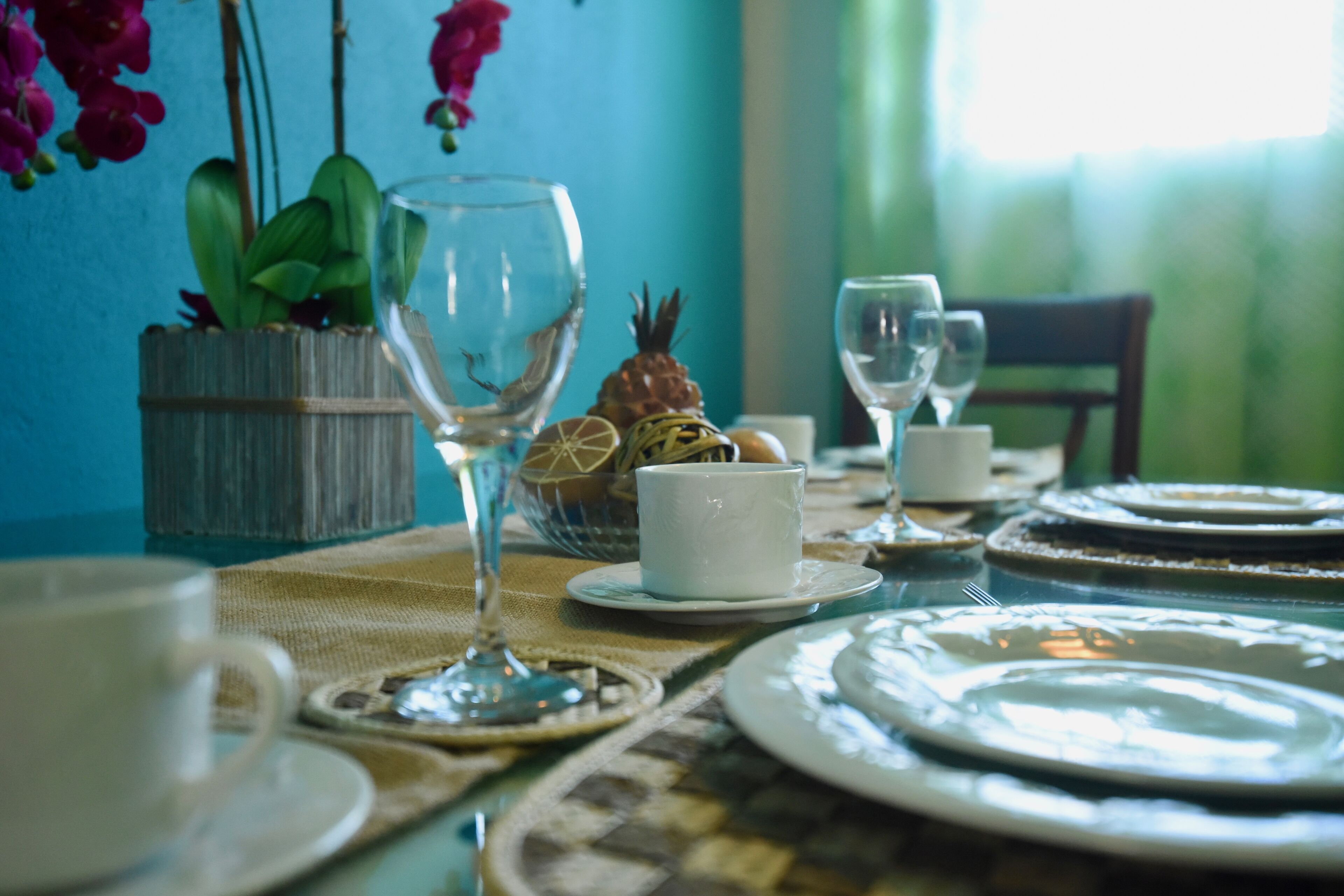Een eettafel gedekt met borden, kopjes en een glas. Op tafel staat een potplant en een ananas.