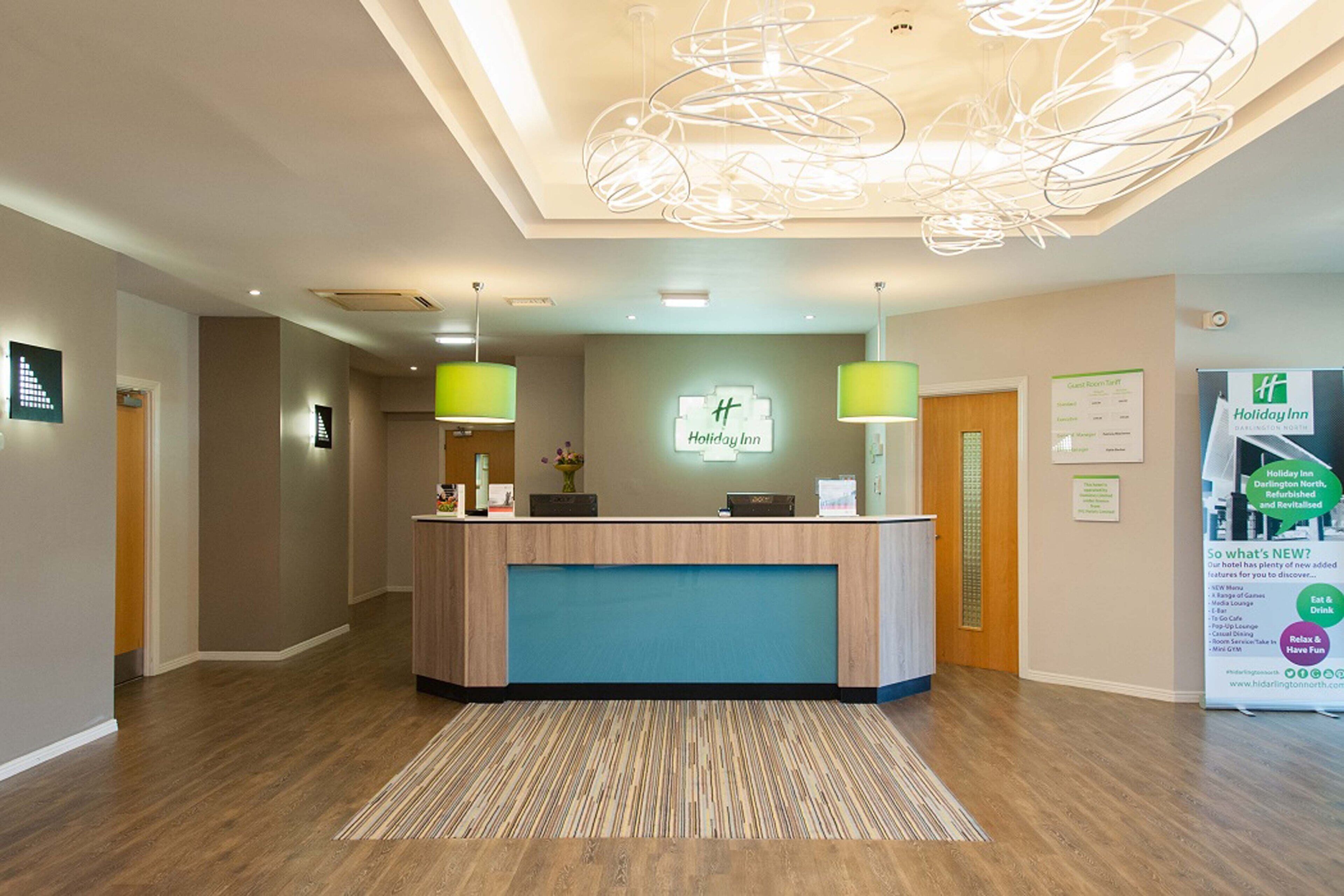 A reception area with a wooden reception desk, two green lamps, a sign reading "Holiday Inn," and a poster advertising new features.