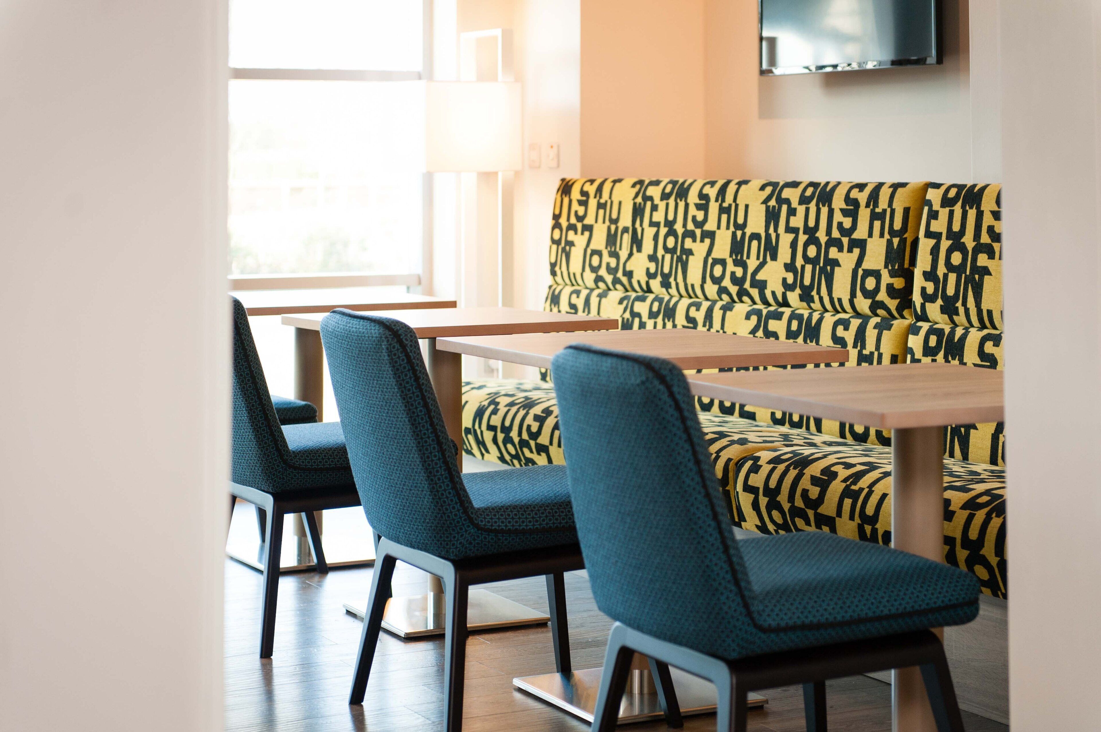 A modern dining area with a wooden table and blue chairs, a yellow patterned sofa, and a TV mounted on the wall.
