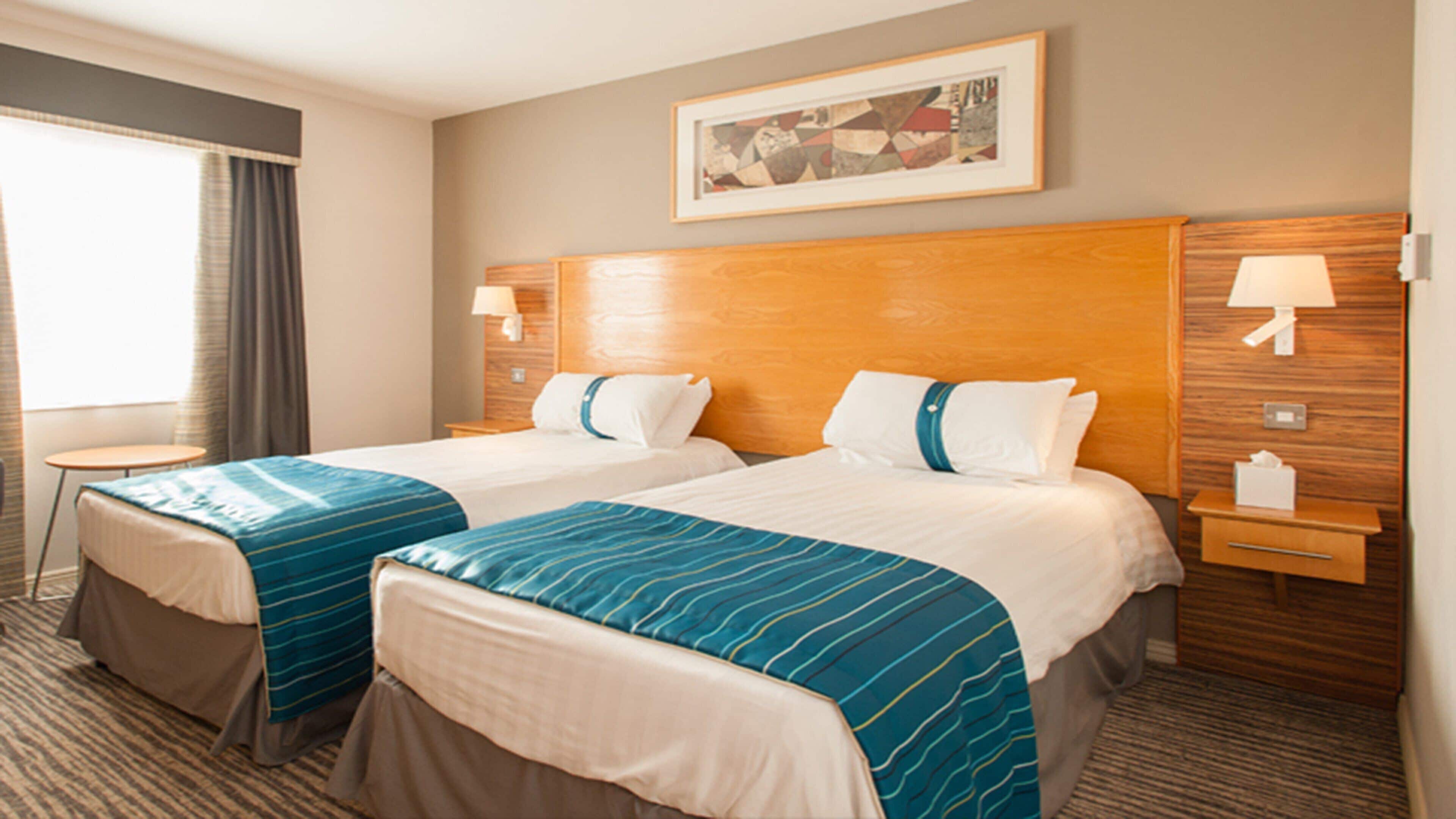 Een hotelkamer met twee bedden, een raam met gordijnen, een schilderij aan de muur en nachtlampjes.
