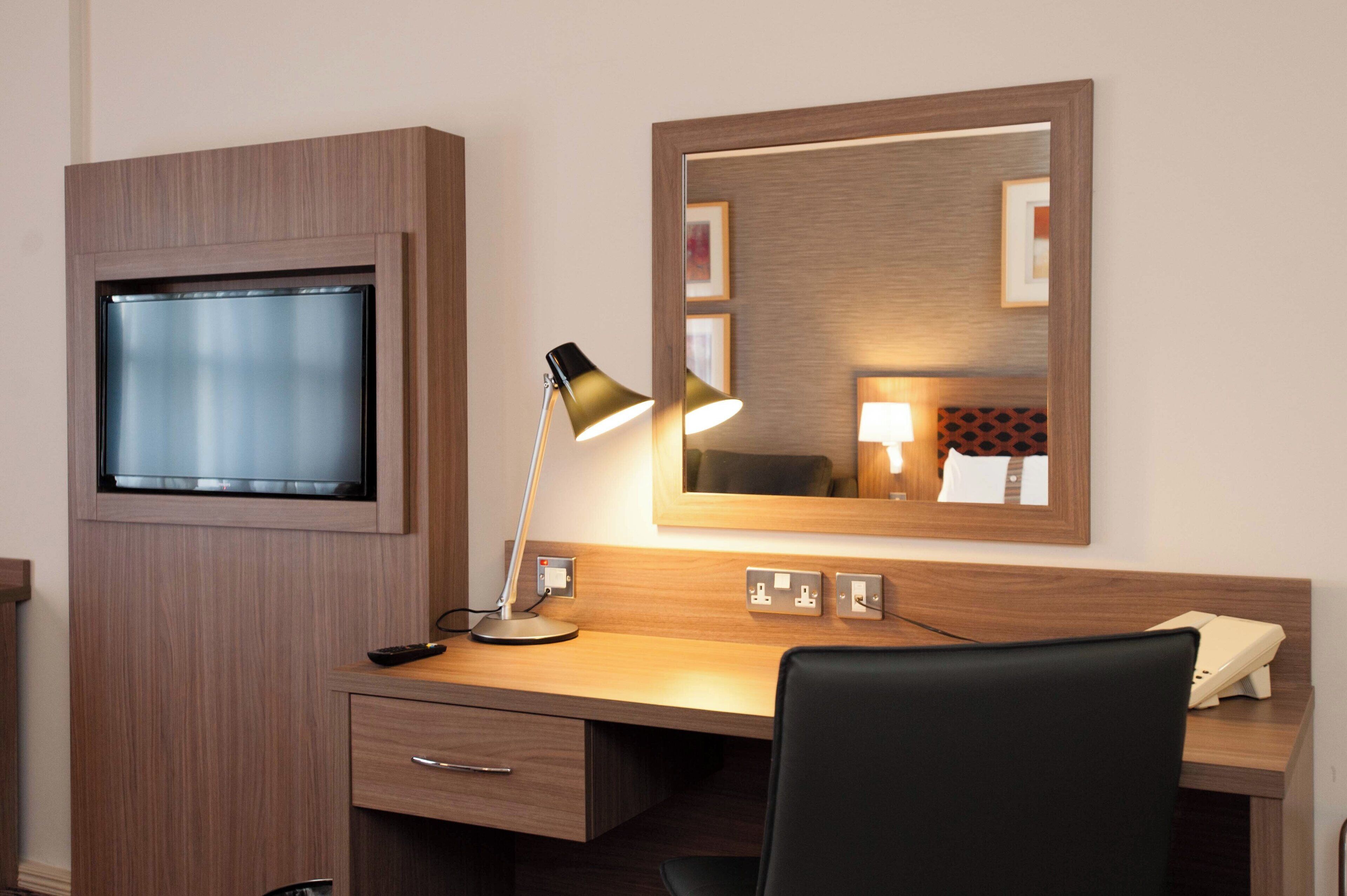 Een hotelkamer met een houten bureau, een televisie, een bureaulamp, een spiegel en een stoel.