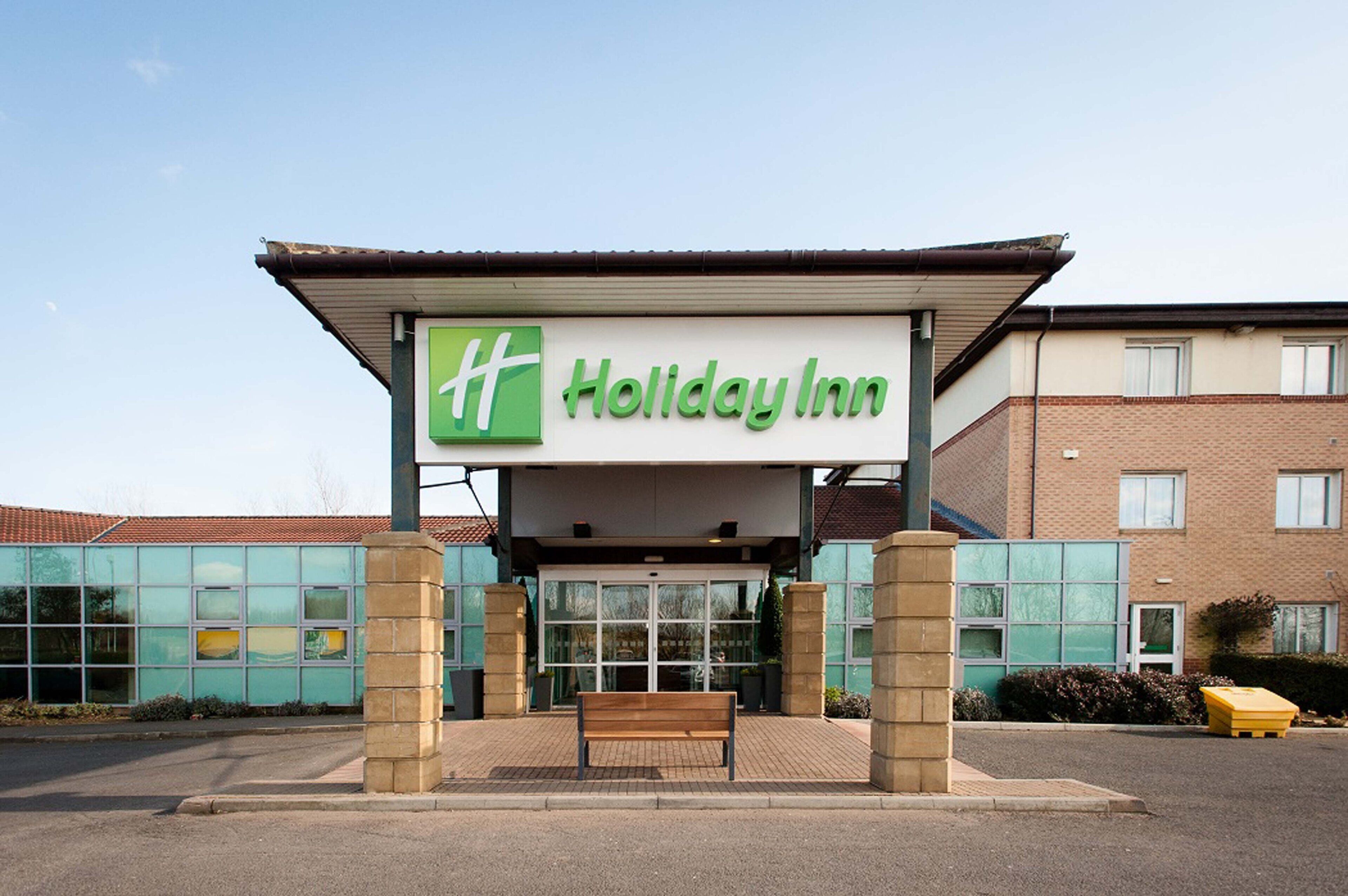 Un hôtel Holiday Inn avec un logo vert, une entrée abritée et un banc devant.