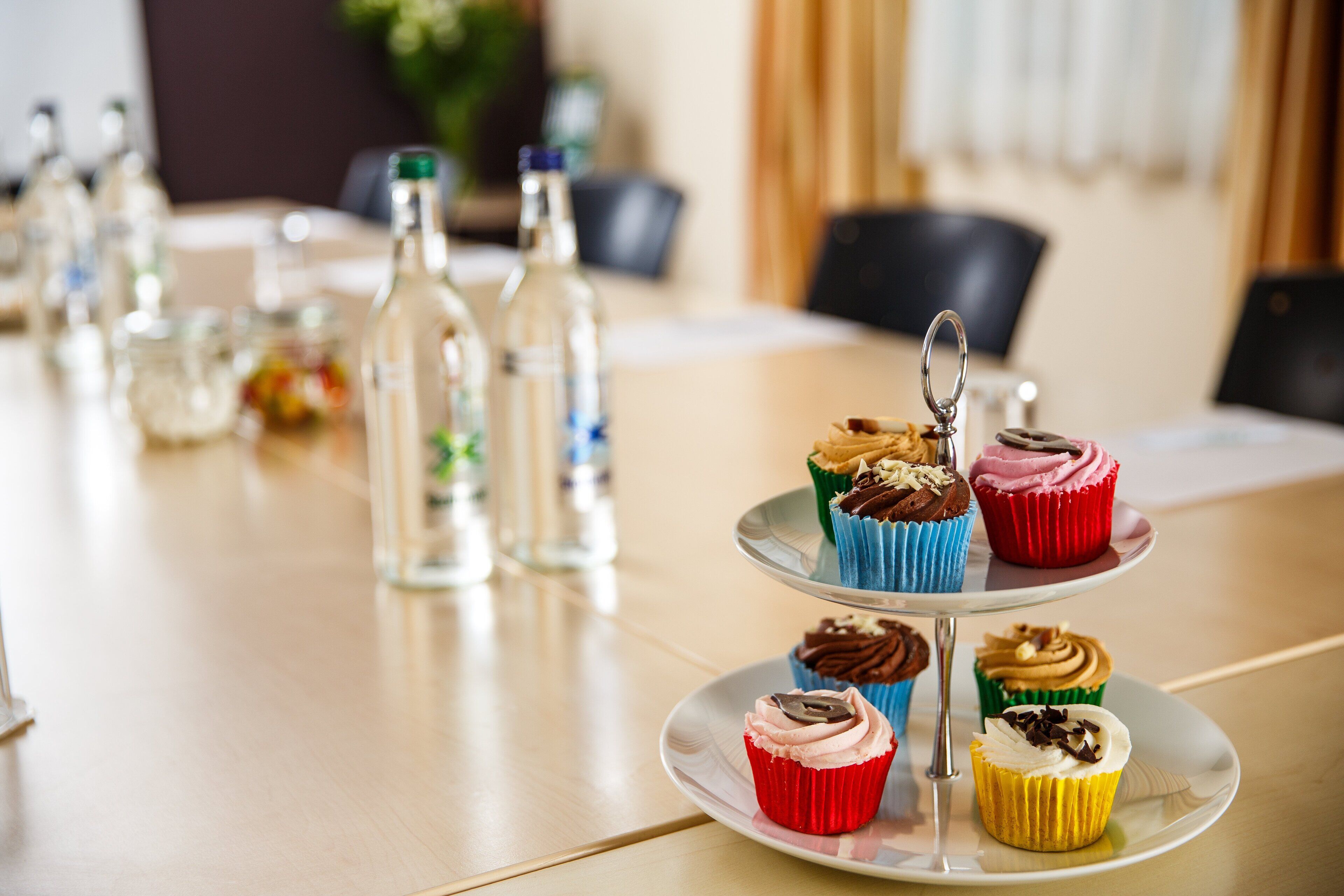 A table with a three-tiered stand holding colorful cupcakes, clear glass bottles, and a small plant.