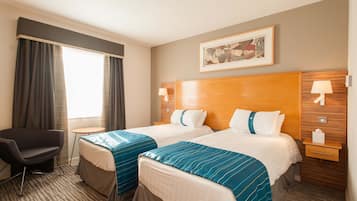 Ein Hotelzimmer mit zwei Betten, einem kleinen Schreibtisch, einem Stuhl, einem Fenster mit Vorhängen und einem gerahmten Bild an der Wand.