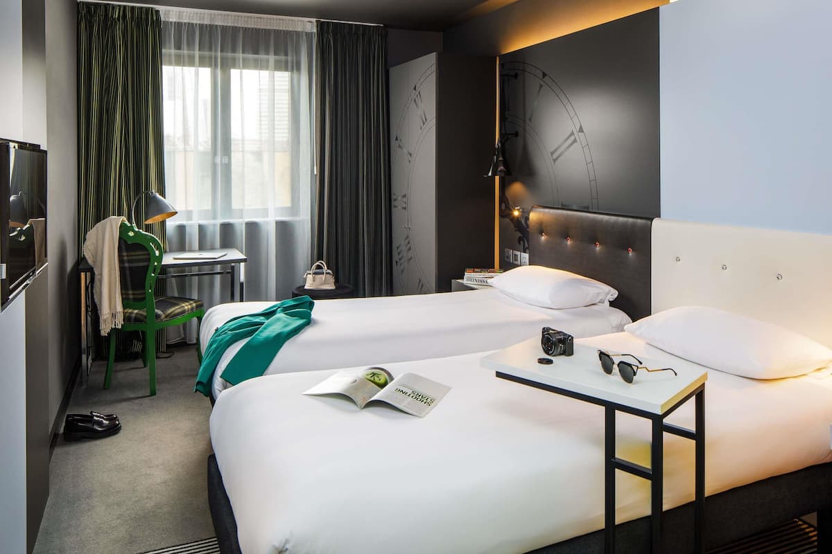 Une chambre d’hôtel avec deux lits, un bureau, une chaise, une lampe et une horloge au mur.