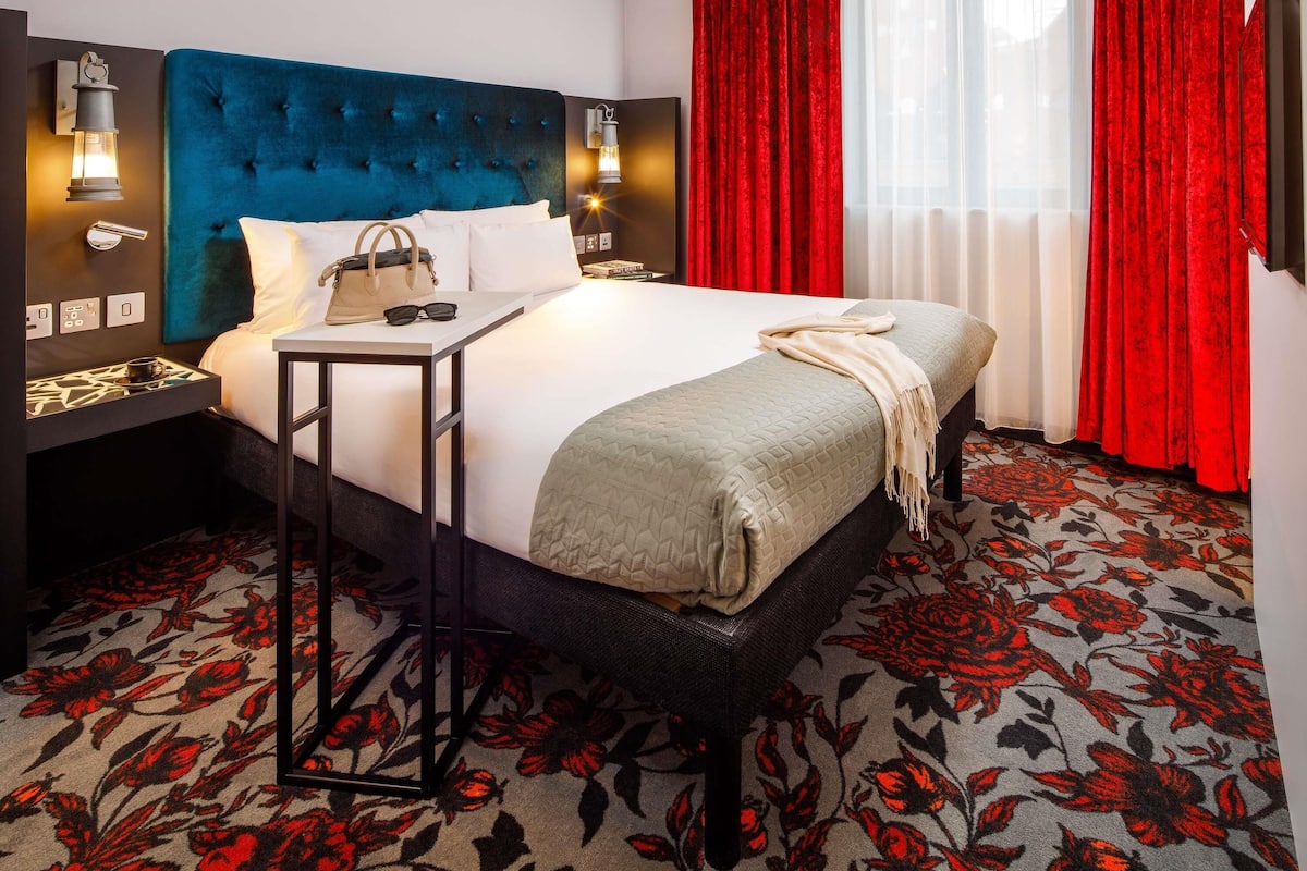 Une chambre d’hôtel avec une tête de lit capitonnée, une table de chevet avec une lampe, un lit recouvert d’une courtepointe et un tapis rouge à motifs floraux.