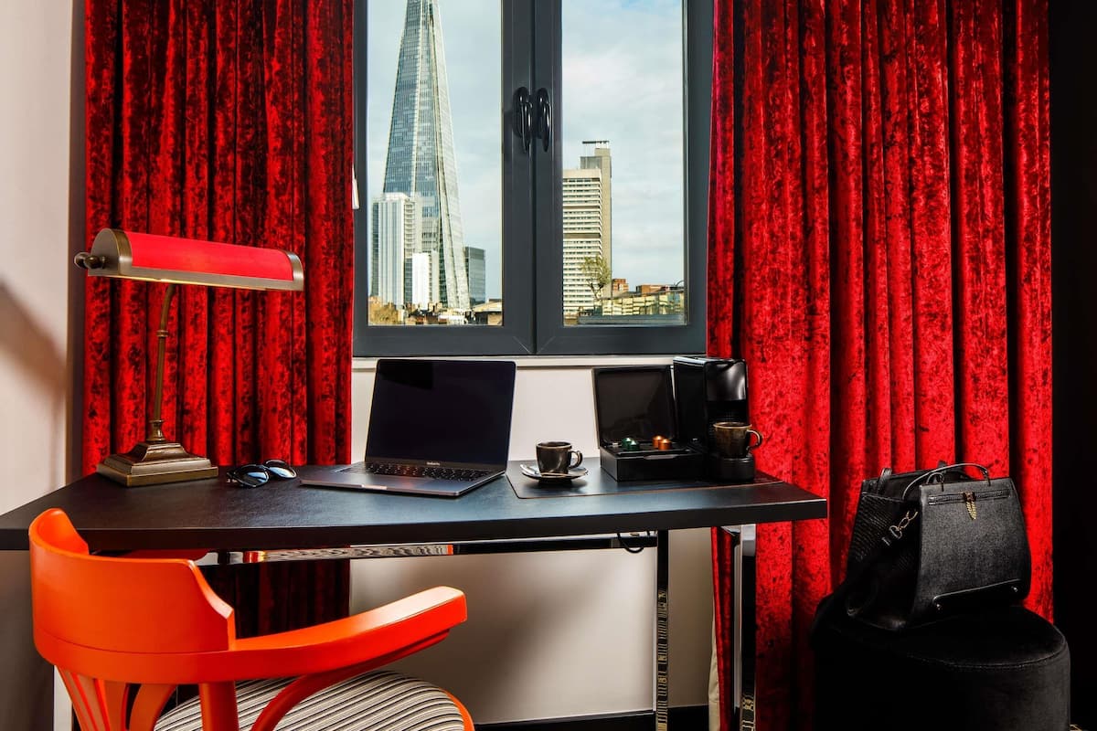 A room with a desk, a laptop, a coffee maker, a red chair, and a view of a tall building through a window.