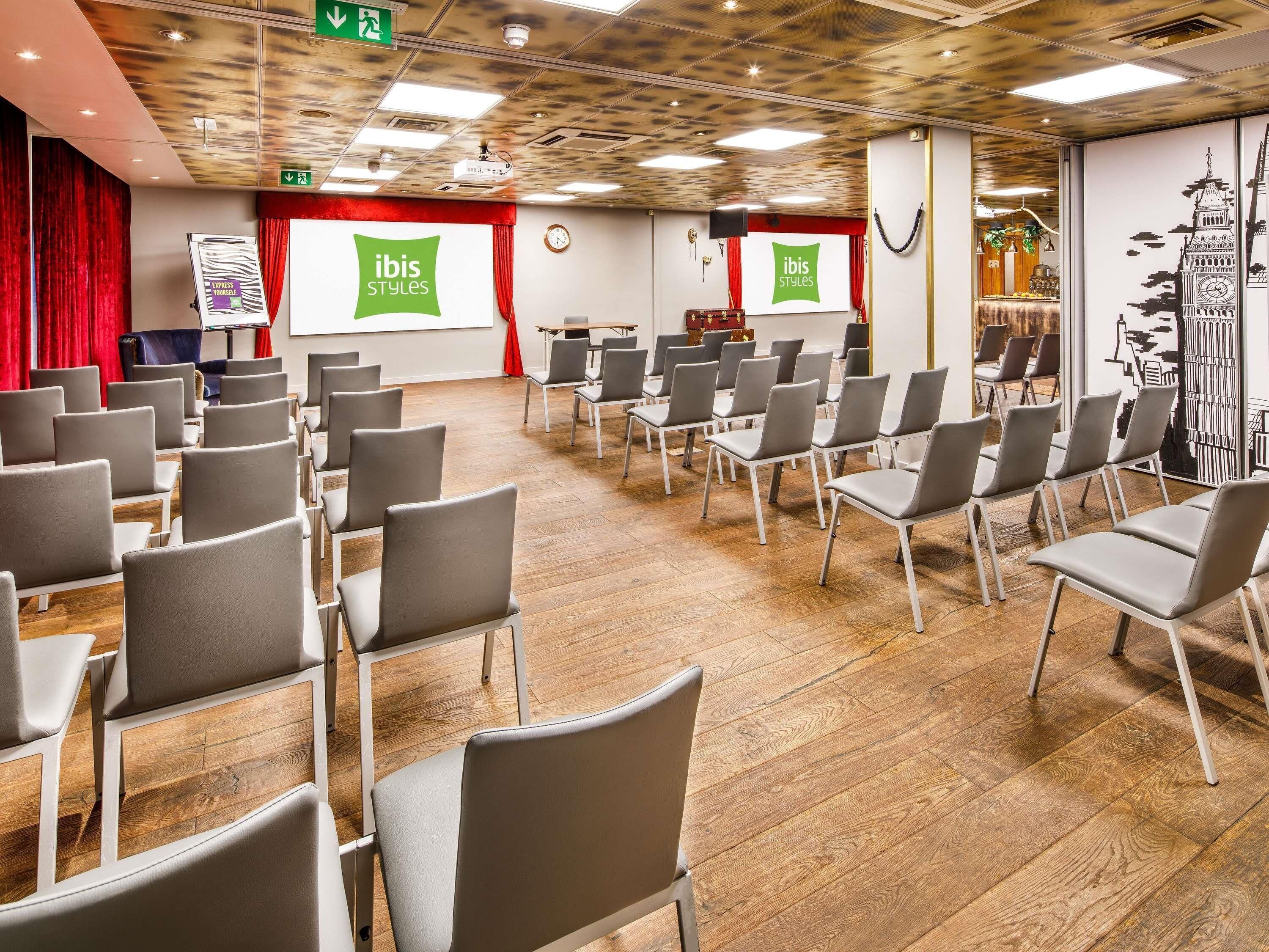 Une salle de conférence avec des rangées de chaises grises, un plancher en bois, un écran de projection et une horloge au mur.