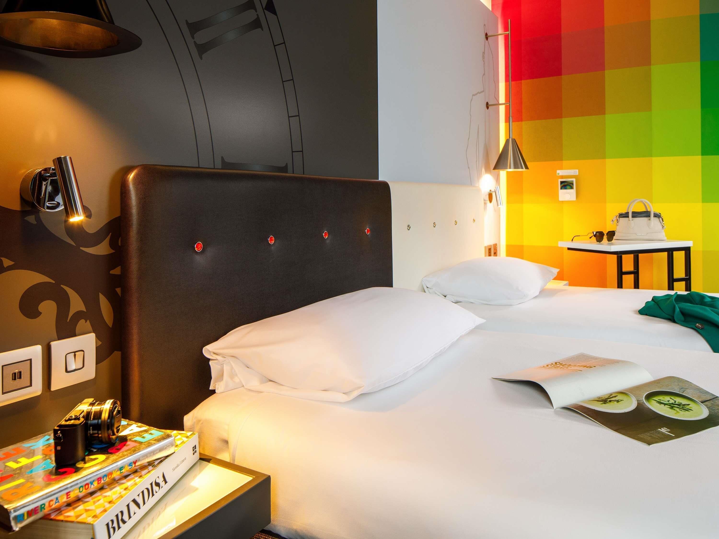 Una habitación de hotel con una cama, una mesita de noche con una cámara y un libro, una mesa con un bolso de mano y gafas de sol, y una pared colorida.