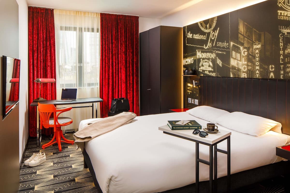 A hotel room with a bed, a desk, a chair, a laptop, a book, and a pair of glasses.