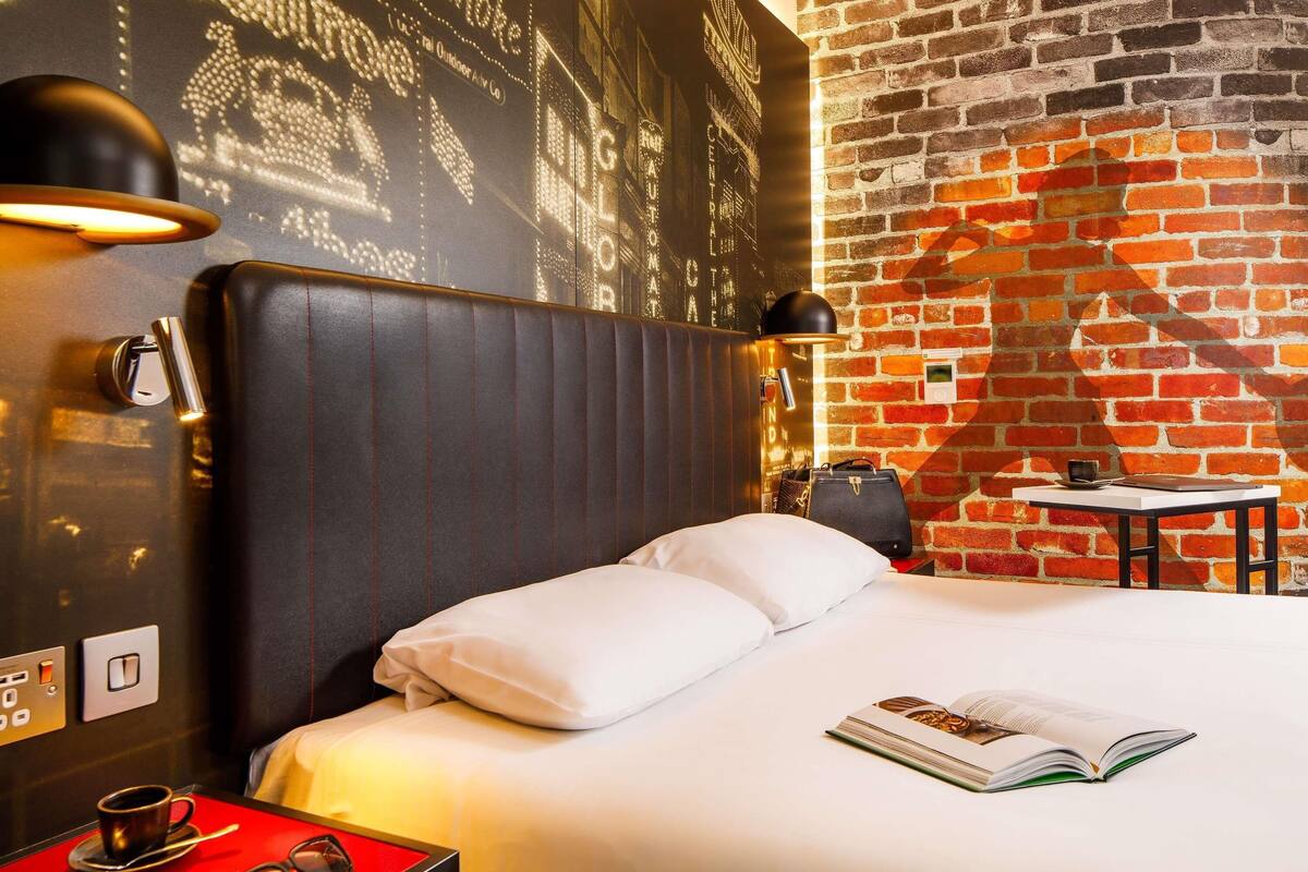 Une chambre d’hôtel avec un mur en briques, un lit recouvert de draps blancs, une table de chevet rouge avec une tasse et des lunettes de soleil, et une petite table avec un livre et une lampe.
