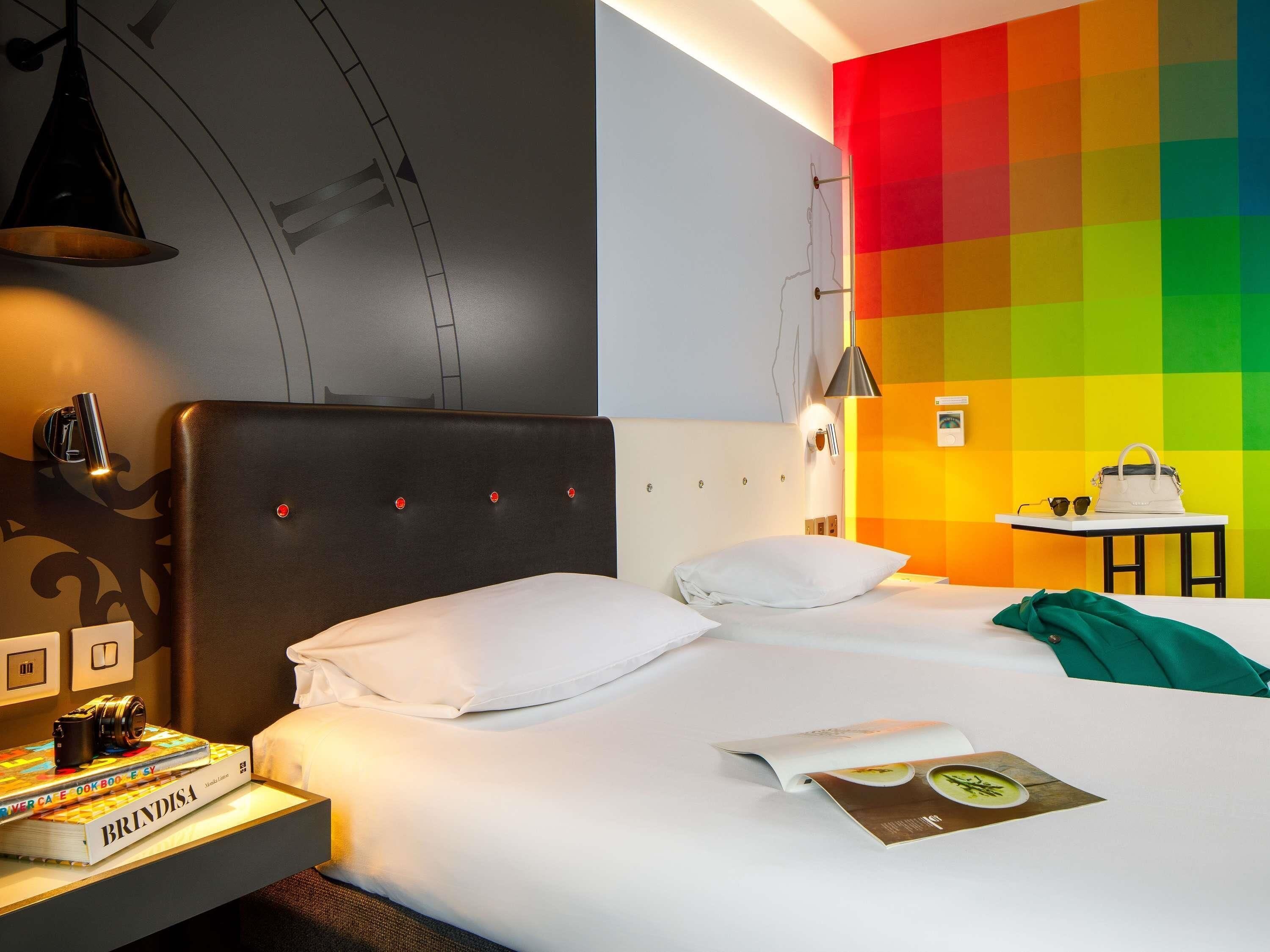 Quarto de hotel com uma cama, uma mesa de cabeceira com livros, um abajur, uma mesa com uma bolsa e uma parede com um padrão colorido.