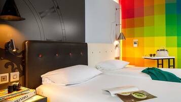 Quarto de hotel com uma cama, uma mesa de cabeceira com livros, um abajur, uma mesa com uma bolsa e uma parede com um padrão colorido.