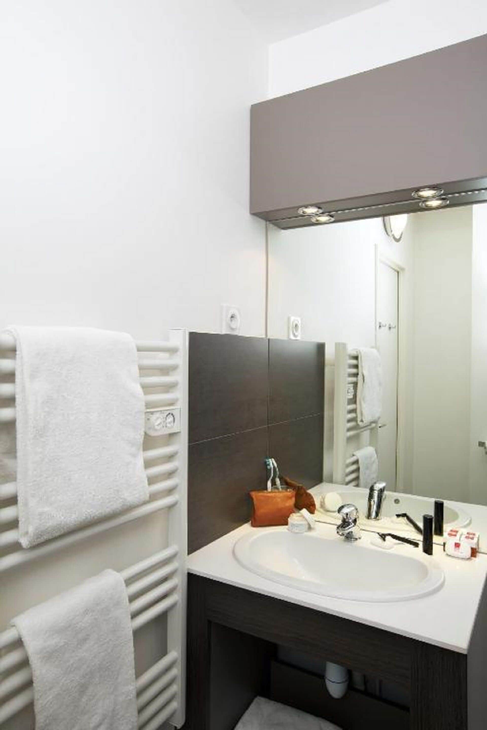 Un baño moderno con un lavamanos blanco, baldosas oscuras, un espejo grande y un toallero con toallas blancas.