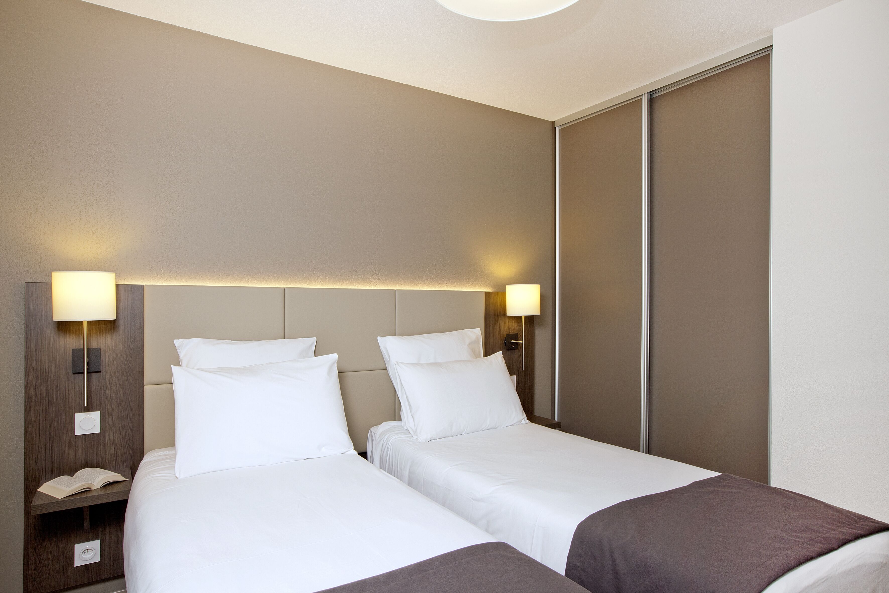 Quarto de hotel com duas camas, uma mesinha de cabeceira com abajur e uma porta de correr.