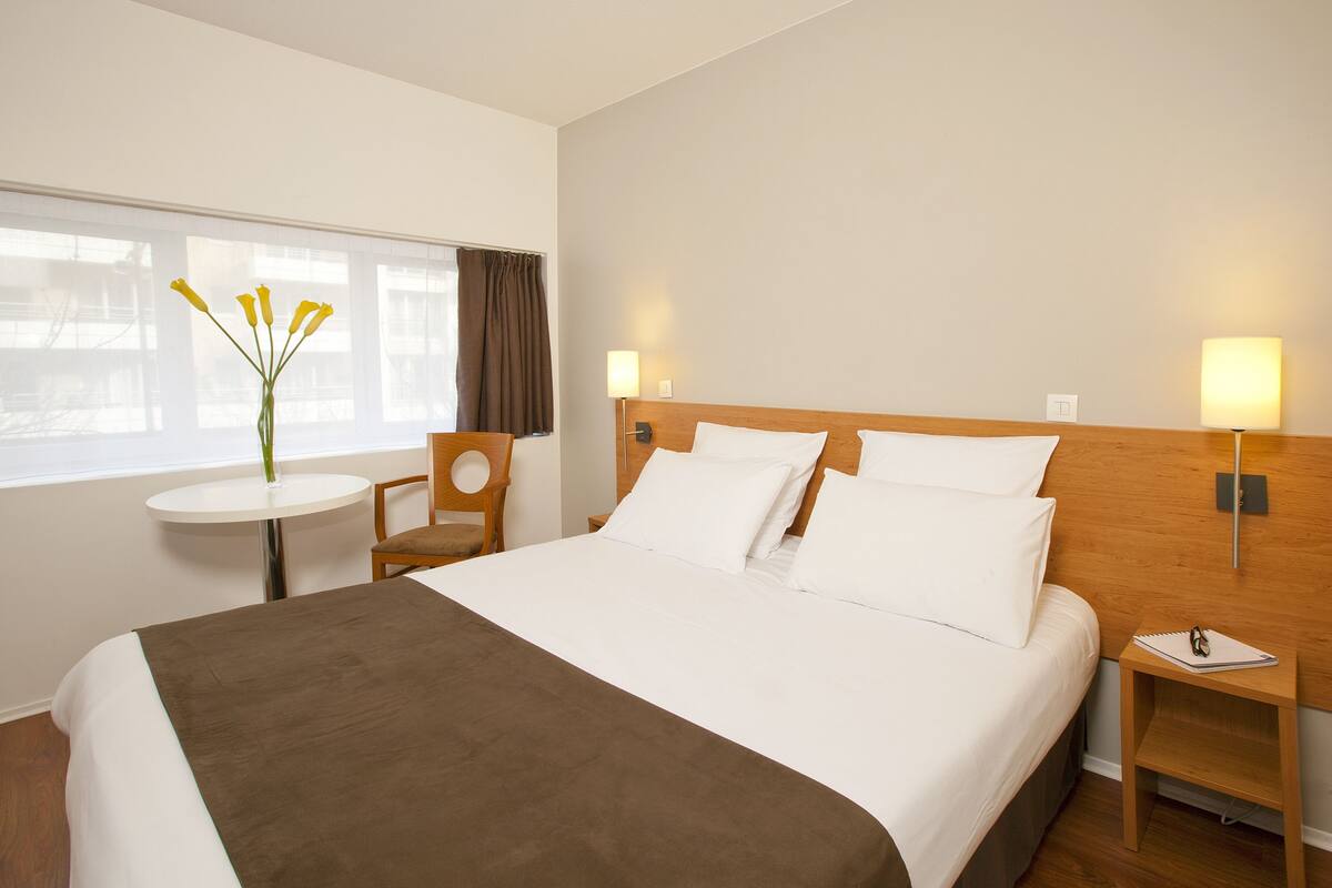 Une chambre d’hôtel avec un grand lit, une petite table ronde, une chaise et un vase rempli de fleurs.