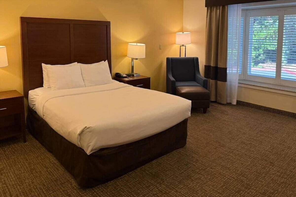 A hotel room with a large bed, bedside lamps, a chair, and a window with curtains.