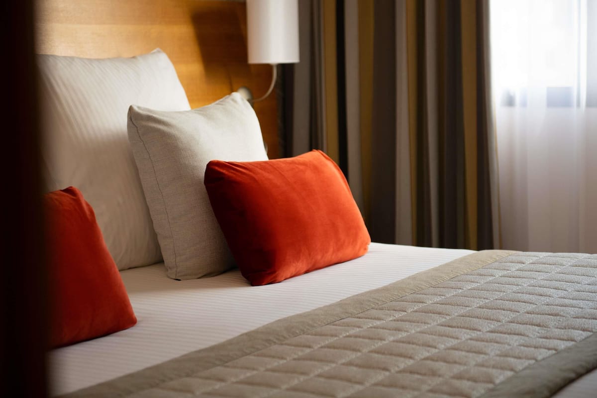 A hotel room with a bed, pillows, a lamp, and curtains.
