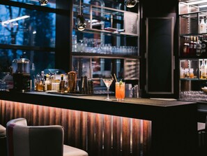 Zona de bar con mostrador de madera, dos vasos de cócteles y una variedad de botellas y vasos al fondo.