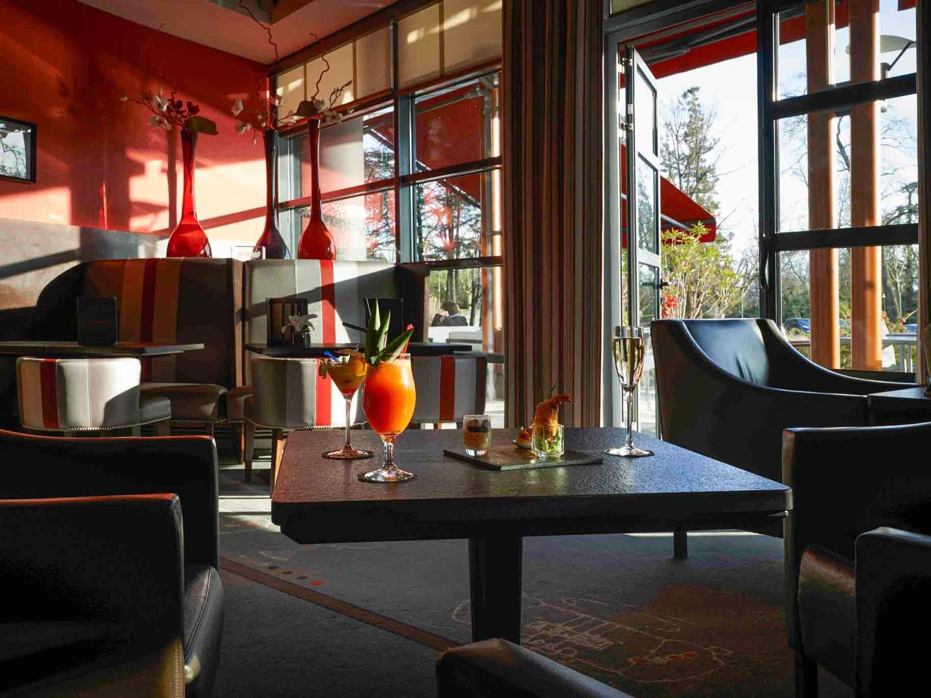 A restaurant with a modern interior, featuring a bar area with a variety of drinks, large windows with a view of the outside, and a mix of seating arrangements including tables and chairs.