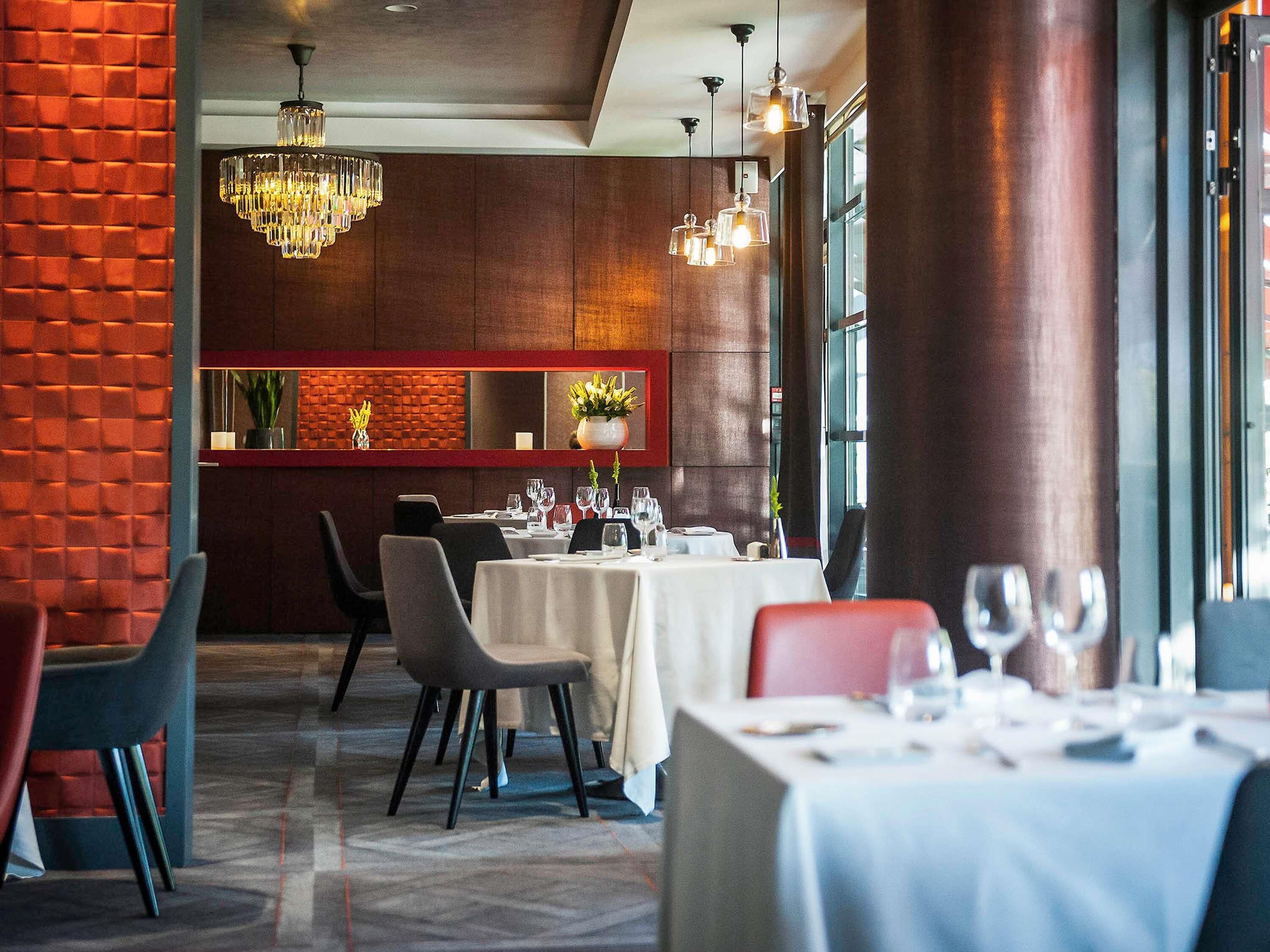 Een restaurant interieur met een bakstenen muur, houten lambrisering en een kroonluchter. Tafels zijn gedekt met witte tafelkleden en glazen.
