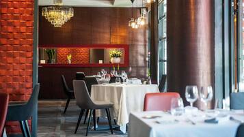 Een restaurant interieur met een bakstenen muur, houten lambrisering en een kroonluchter. Tafels zijn gedekt met witte tafelkleden en glazen.