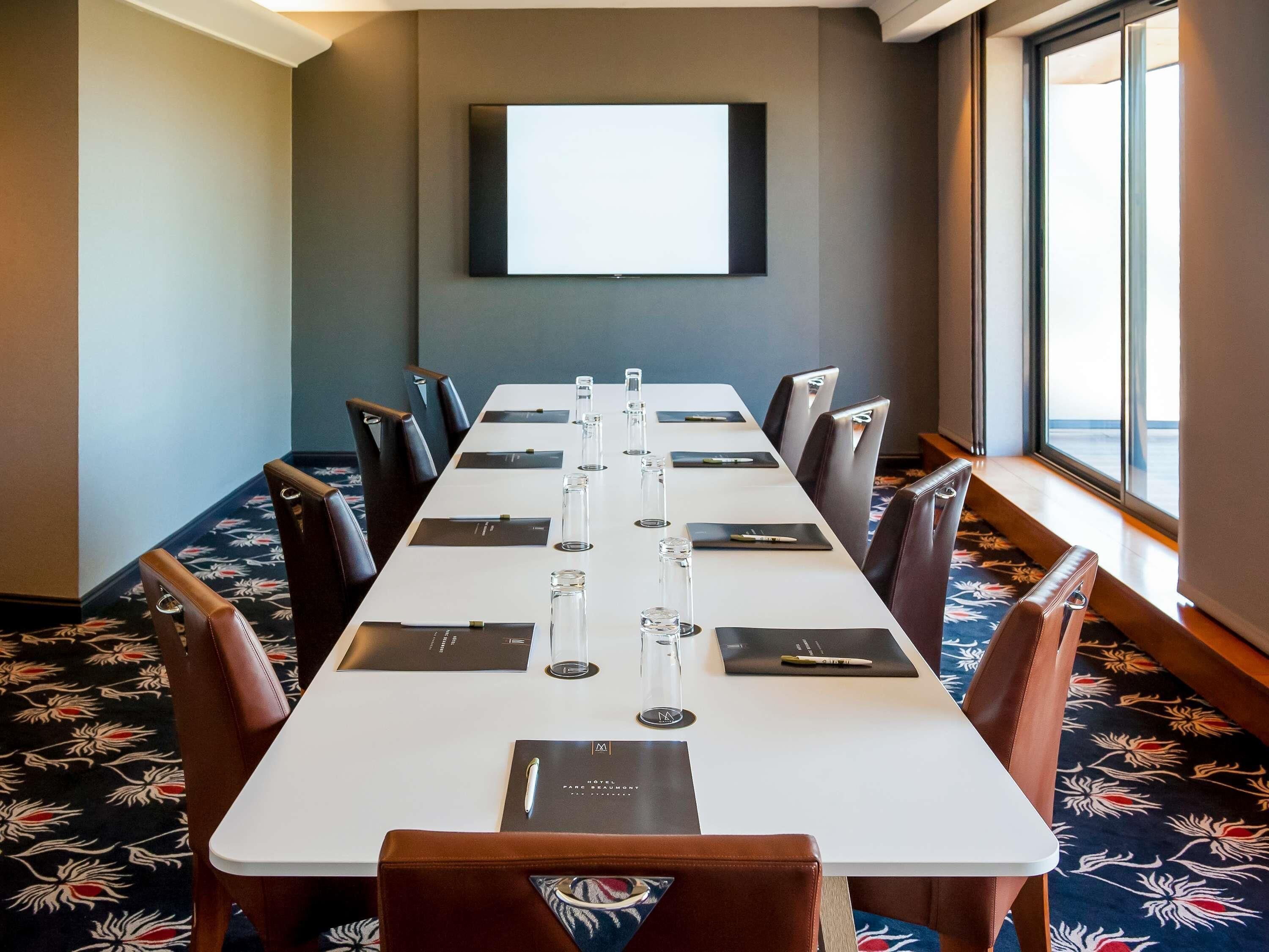 A conference room with a long table, chairs, a projector screen, and a window with a view.