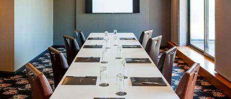 A conference room with a long table, chairs, a projector screen, and a window with a view.
