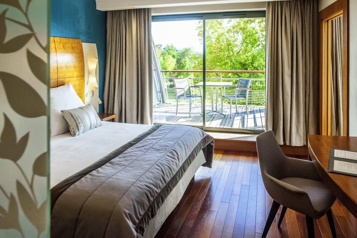 A hotel room with a bed, a desk, a chair, and a balcony with outdoor furniture.