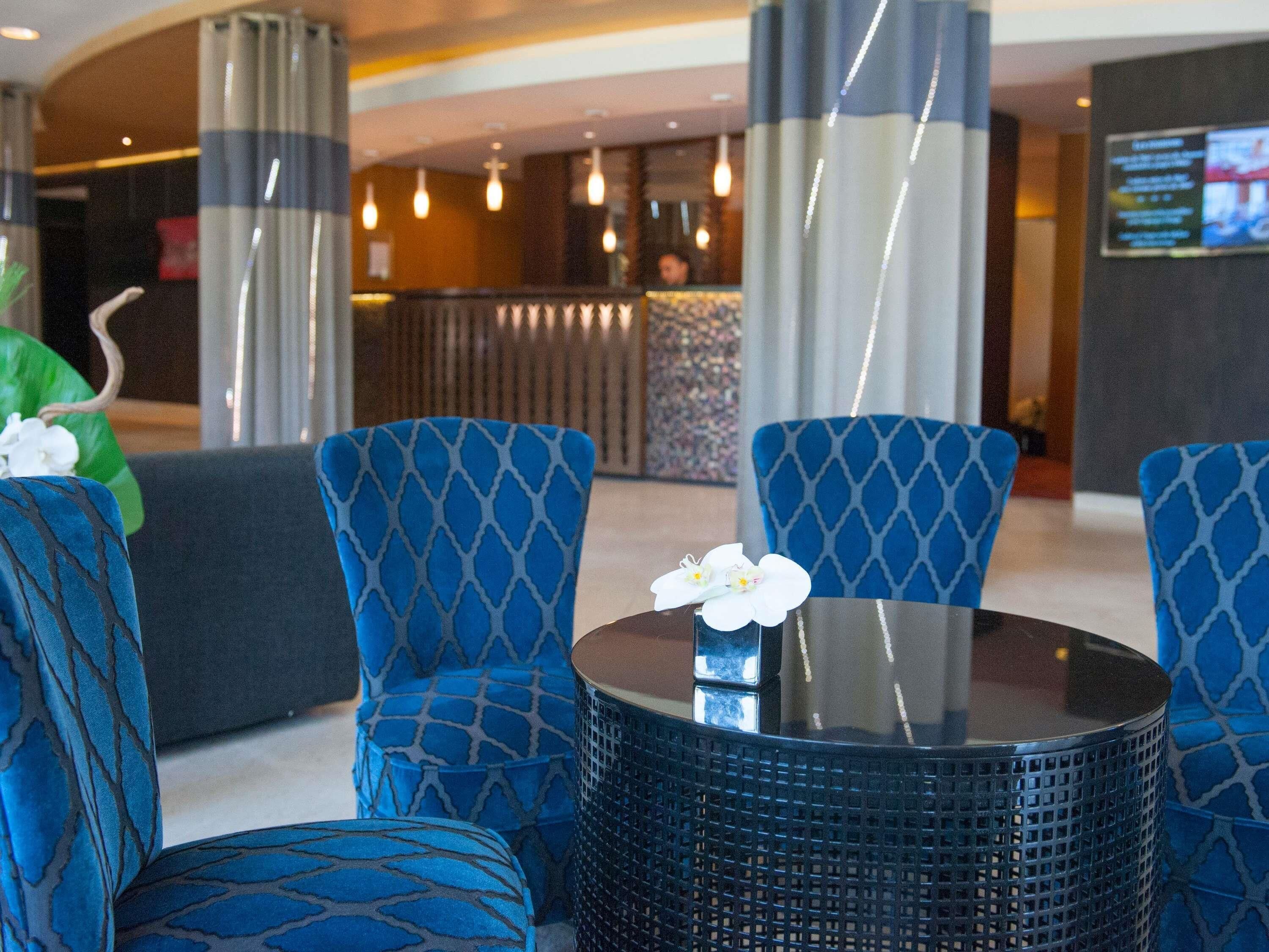 A lobby area with blue patterned chairs, a round table with a vase of white flowers, and a reception desk in the background.