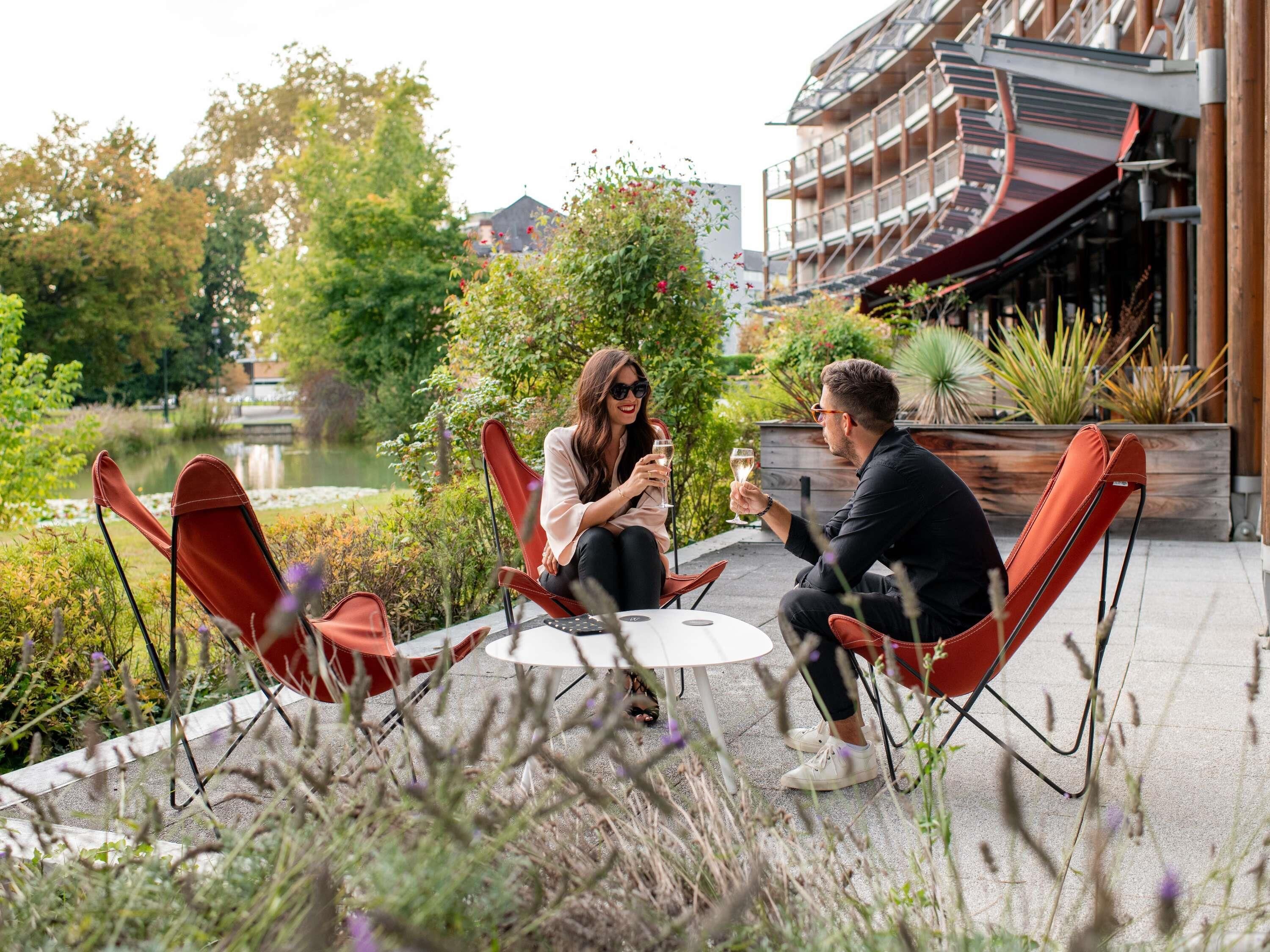 Una pareja sentada en una mesa al aire libre con sillas, rodeada de vegetación y un edificio al fondo.