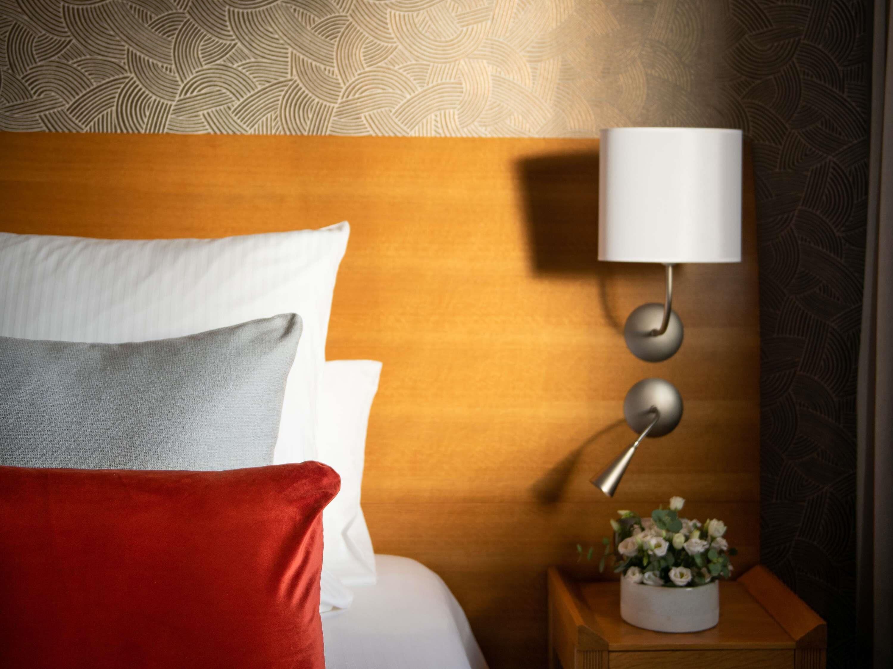 Un lit bien fait, avec des oreillers blancs et gris, un oreiller décoratif rouge et une tête de lit en bois. On y trouve une table de chevet avec une lampe blanche et un vase de fleurs.
