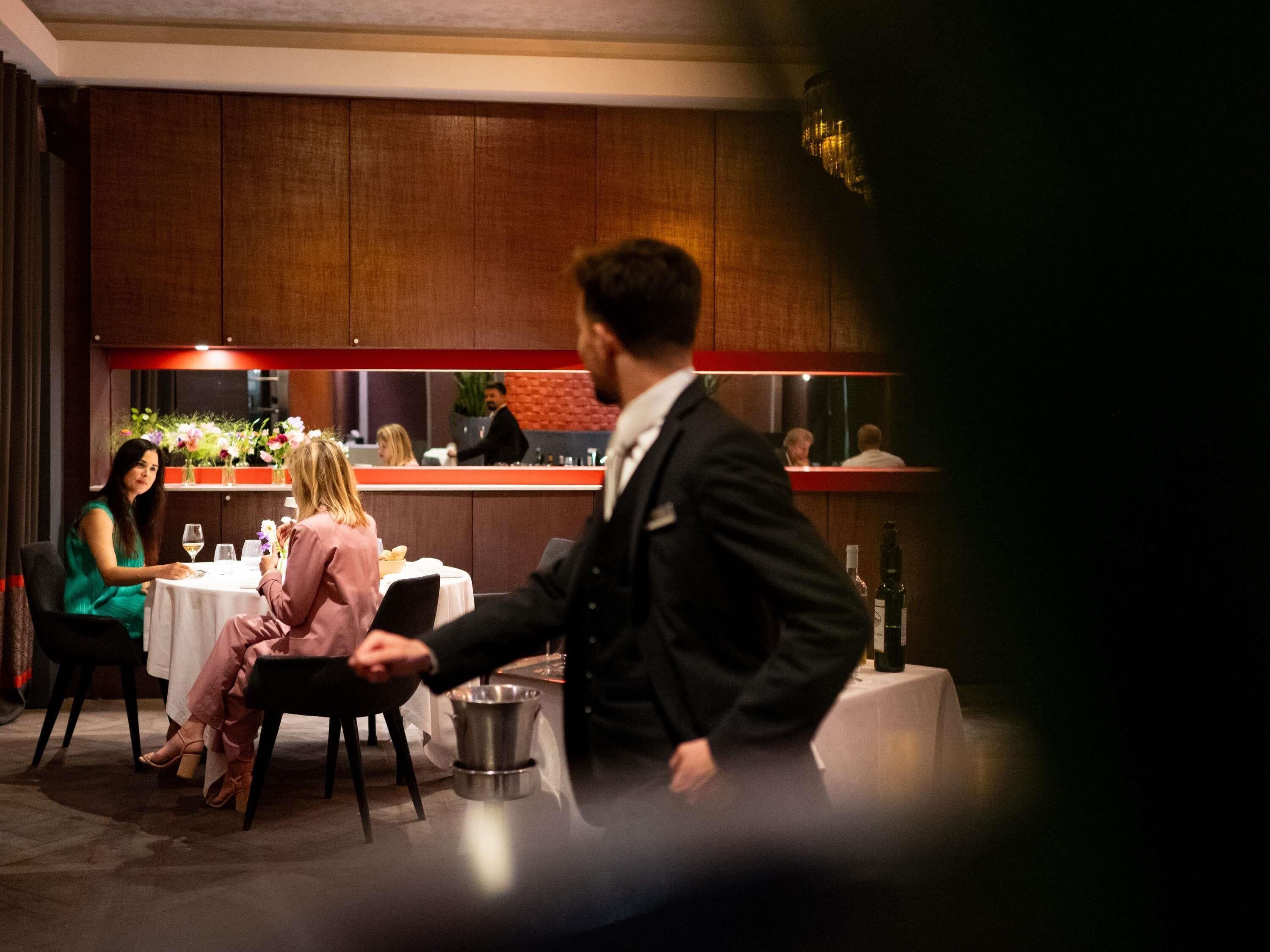 A restaurant interior with wooden paneling, a bar area, and a waiter serving a table.