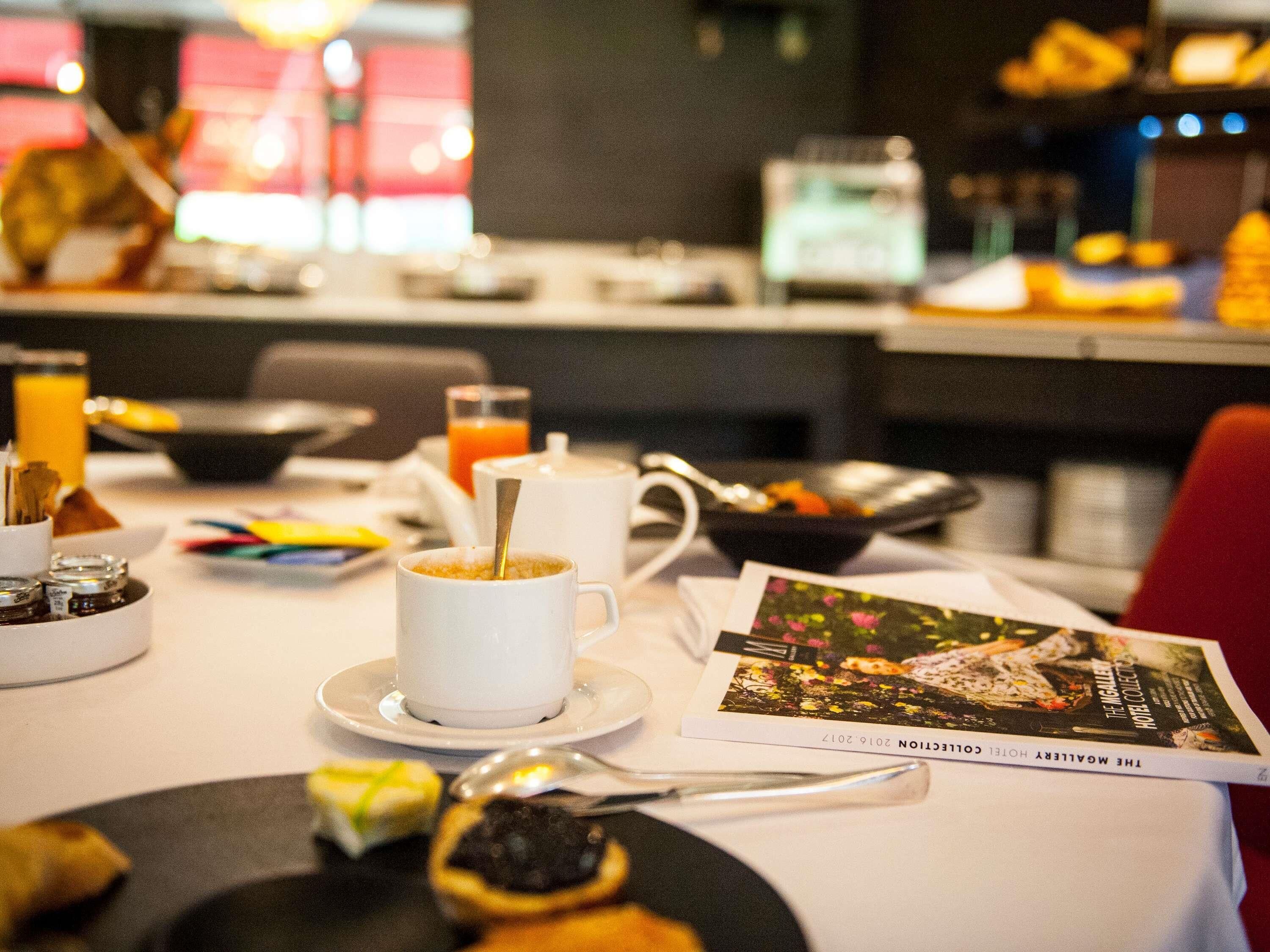 Une table de petit-déjeuner avec une tasse de café, un verre de jus d’orange, un bol de fruits et un magazine.