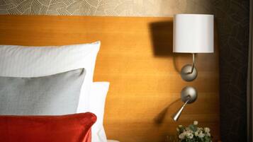 A hotel room with a wooden headboard, a white pillow, a grey pillow, a red throw blanket, a wall-mounted lamp, and a floral arrangement.