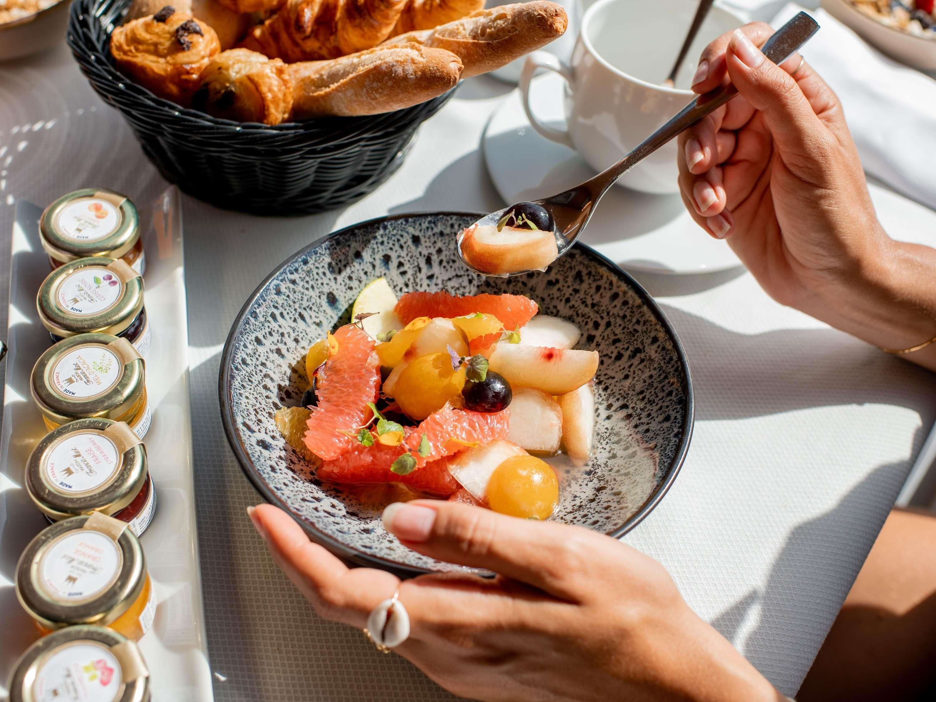 A person holding a spoon with food, a basket of bread, and jars of jam on a table.