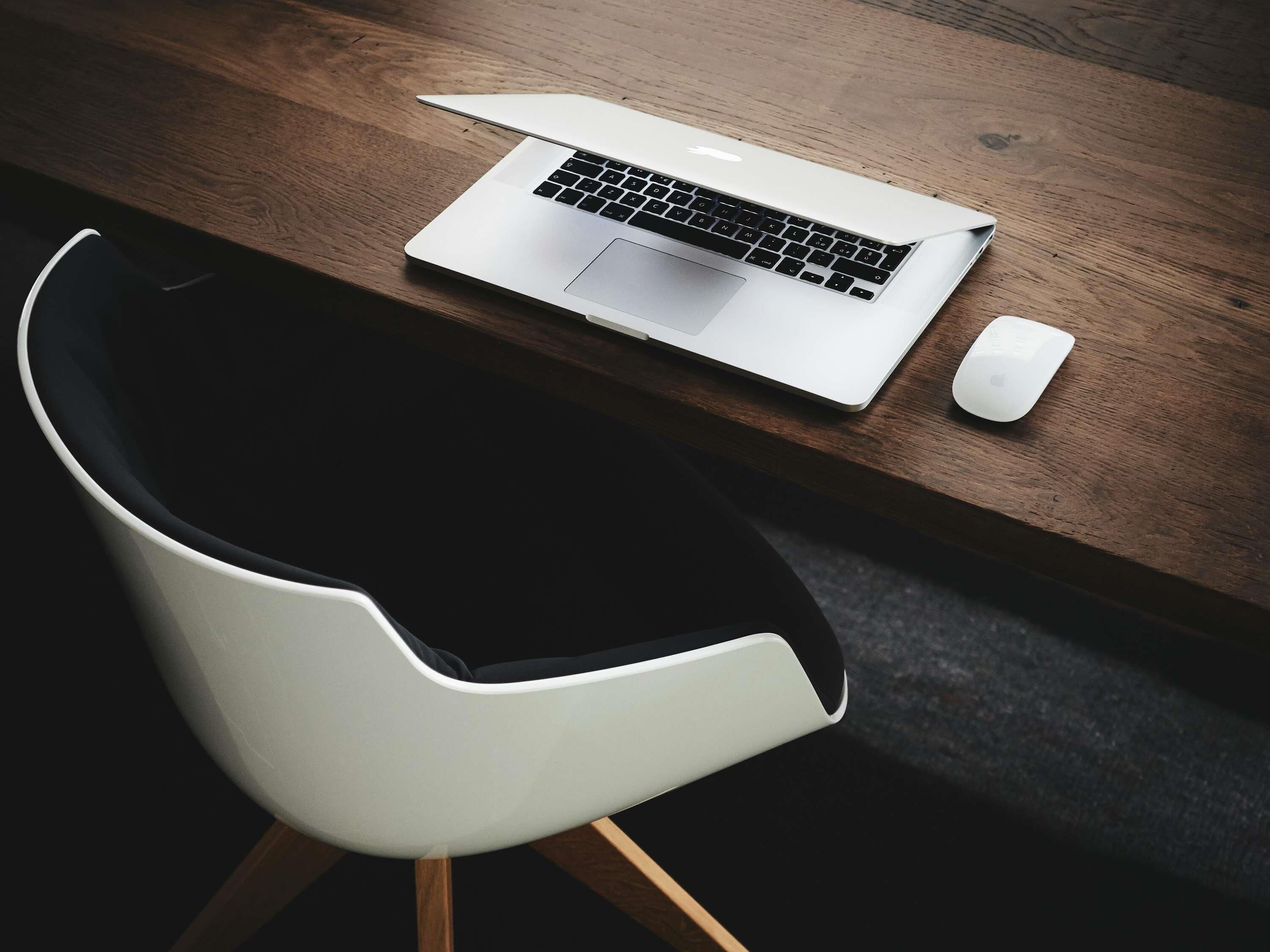 A laptop on a wooden desk with a white chair and a mouse.