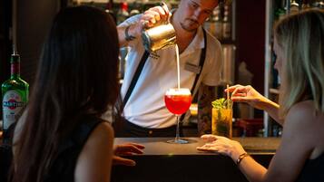 Une scène de bar : un barman verse une boisson dans un verre, une bouteille de Martini est posée sur le comptoir, et un cocktail avec une paille est visible.