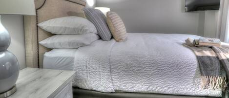Uma cama bem arrumada com travesseiros e um cobertor, um abajur e uma TV de tela plana na parede.