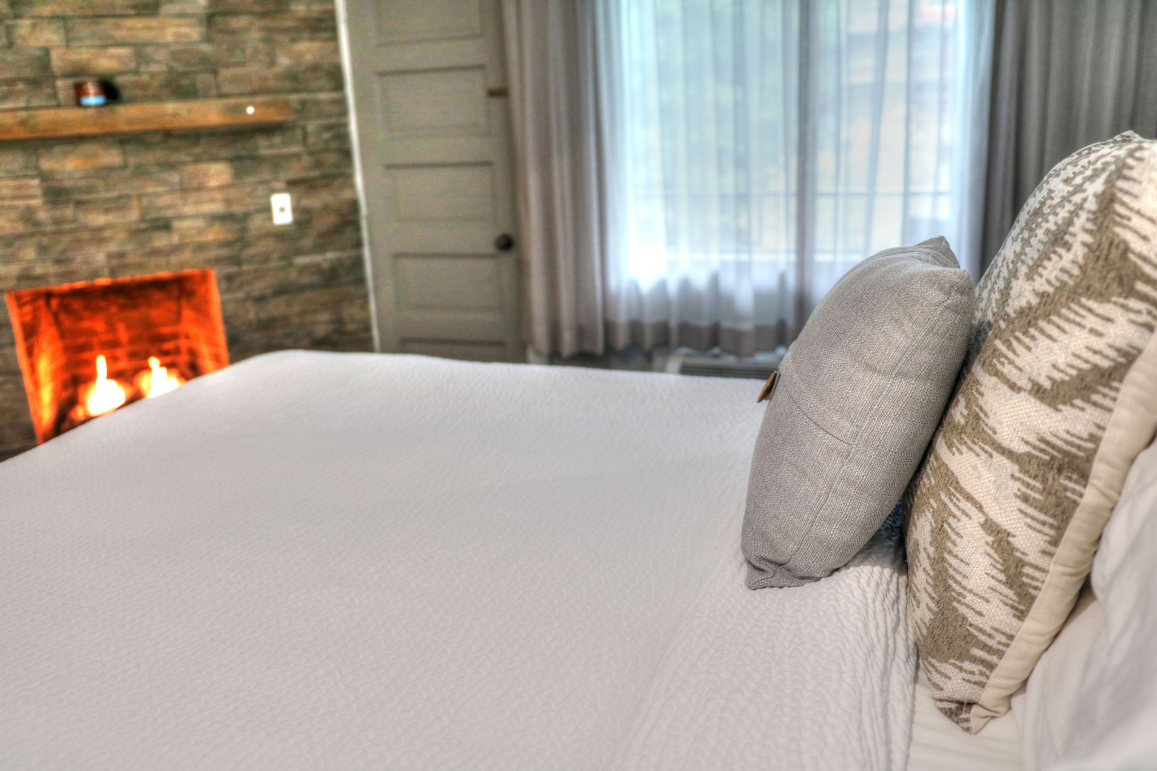 Una cama bien hecha con almohadas, una pared de piedra y una chimenea con llamas visibles.