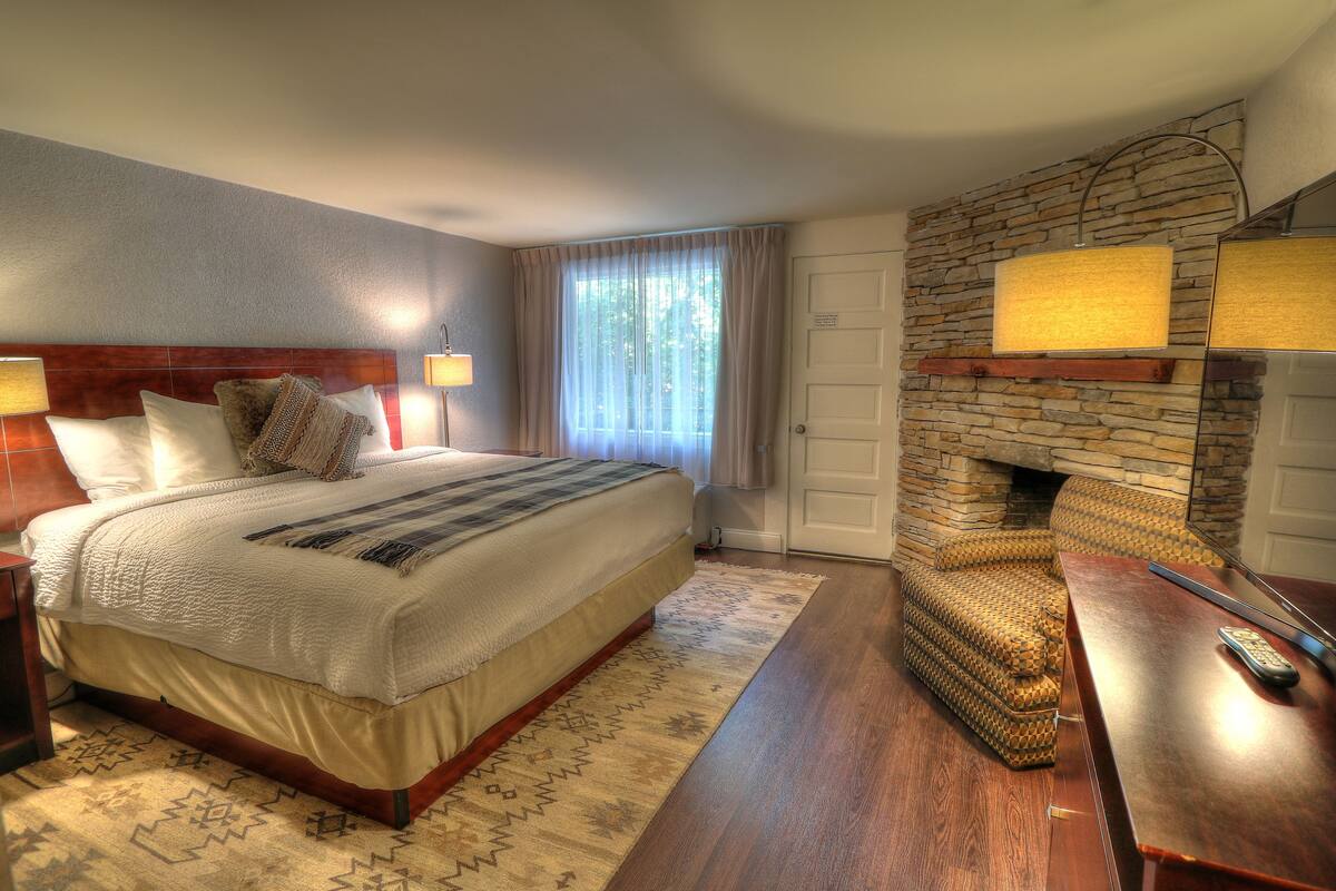Quarto de hotel com uma cama grande, lareira de pedra, escrivaninha de madeira e janela com cortinas.