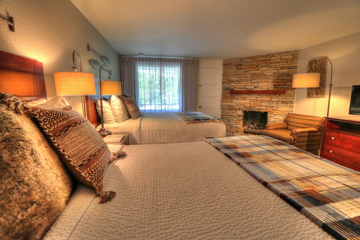 Quarto de hotel com duas camas, parede de pedra, lareira e uma janela grande com cortinas.