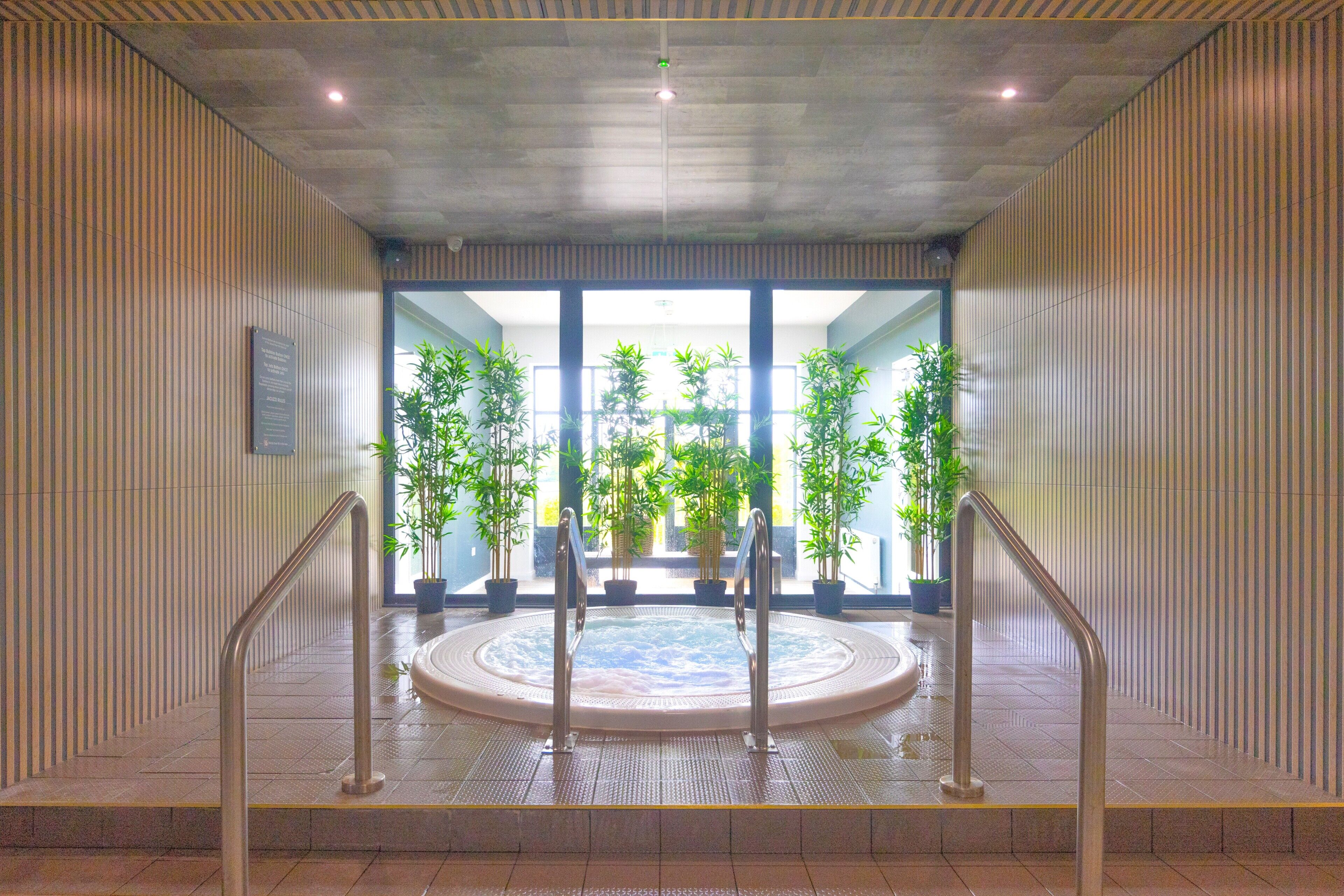 A modern indoor pool area with a central hot tub, surrounded by potted plants and metal railings.